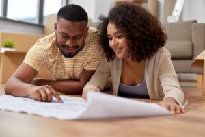 mortgage, people and real estate concept - happy african american couple with boxes and blueprint moving to new home
