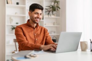 Portrait Of Smiling Middle Eastern Male Manager Working On Laptop Computer In Modern Office, Cheerful Guy Sitting At Desk And Using Pc, Looking At Screen Typing On Keyboard, Enjoying Remote Job