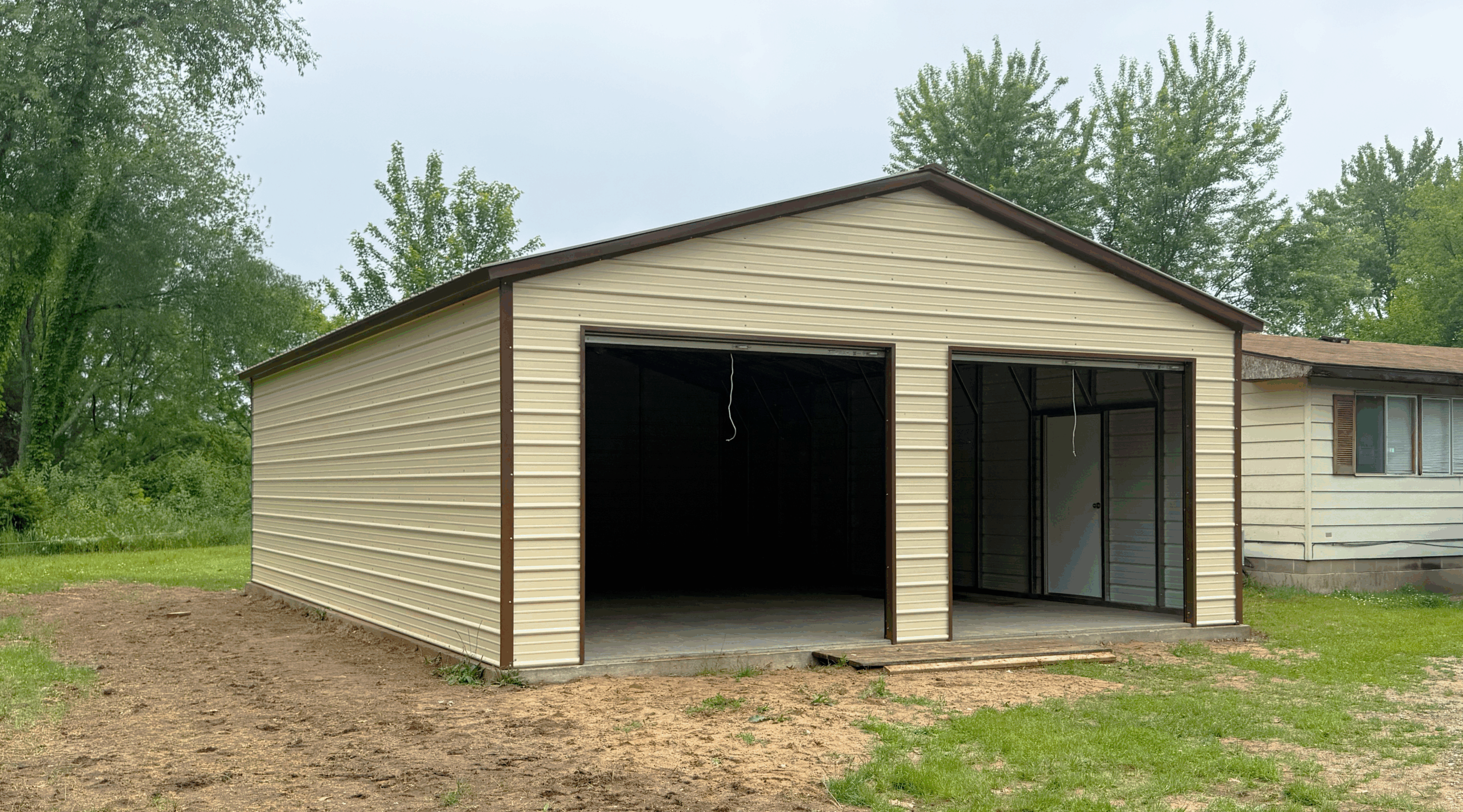 This two-car metal garage, in pebble beige with earth brown trim, features white roll-up doors and a side walk-in door. It was expertly installed by American Steel Carports, Inc. in Albany, KY.