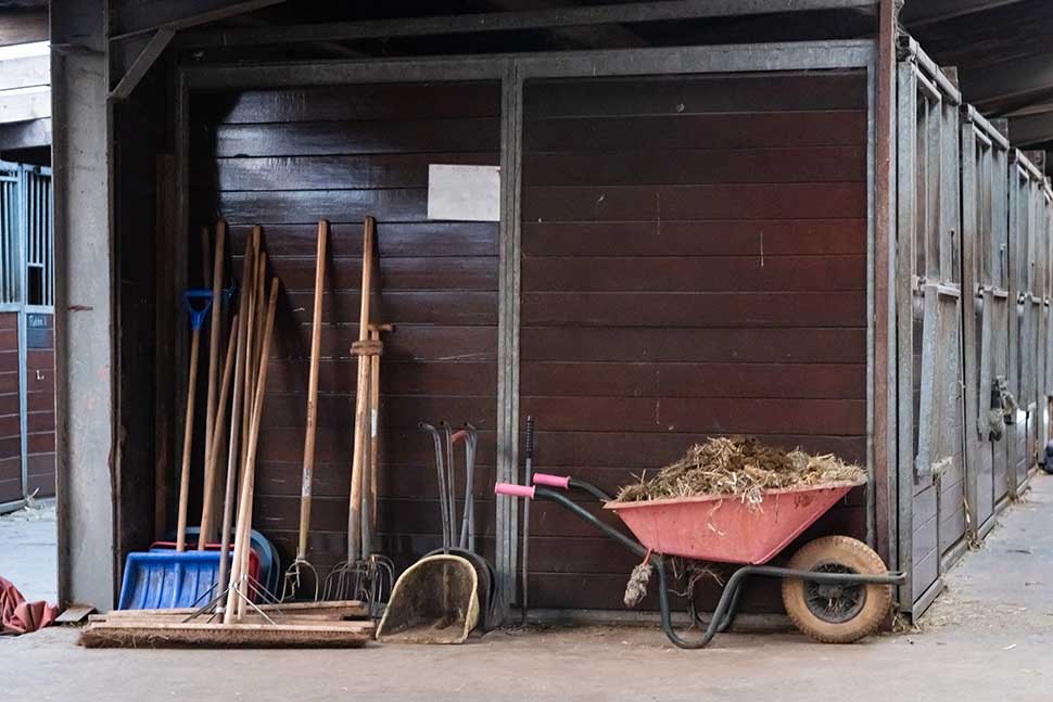 A wheelbarrow, rake, and various tools are arranged in a stable, highlighting essential fall maintenance for Oklahoma steel barns.